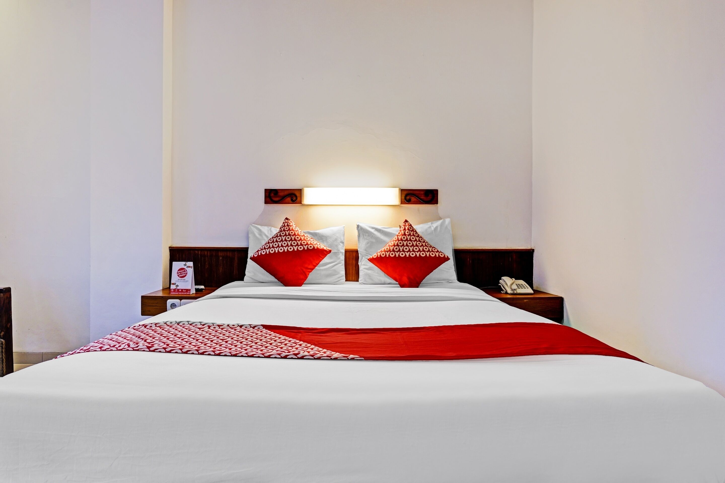A neatly made bed with white and red bedding, a bedside table with a lamp, and a wall-mounted light above the bed.
