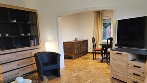 Una sala de estar con un sofá, una mesa de centro de vidrio, un televisor, un mueble de madera y una estantería.