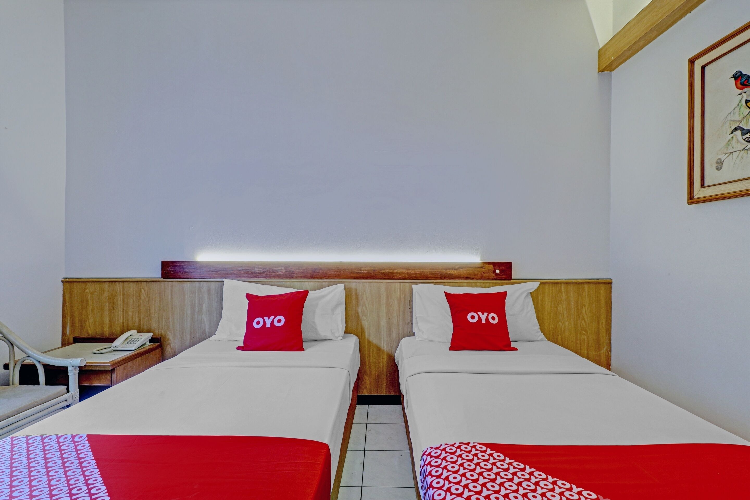 A hotel room with two beds, each with a red pillow labeled "OYO," a wooden headboard, a bedside table with a phone, and a framed picture on the wall.