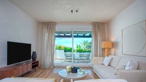 A modern living room with a flat-screen TV, a round coffee table, and a sofa. There is a large window with curtains, a lamp, and a view of greenery outside. - Exclusive Loft With Private Terrace in Marbella. Incosol II (Marbella)