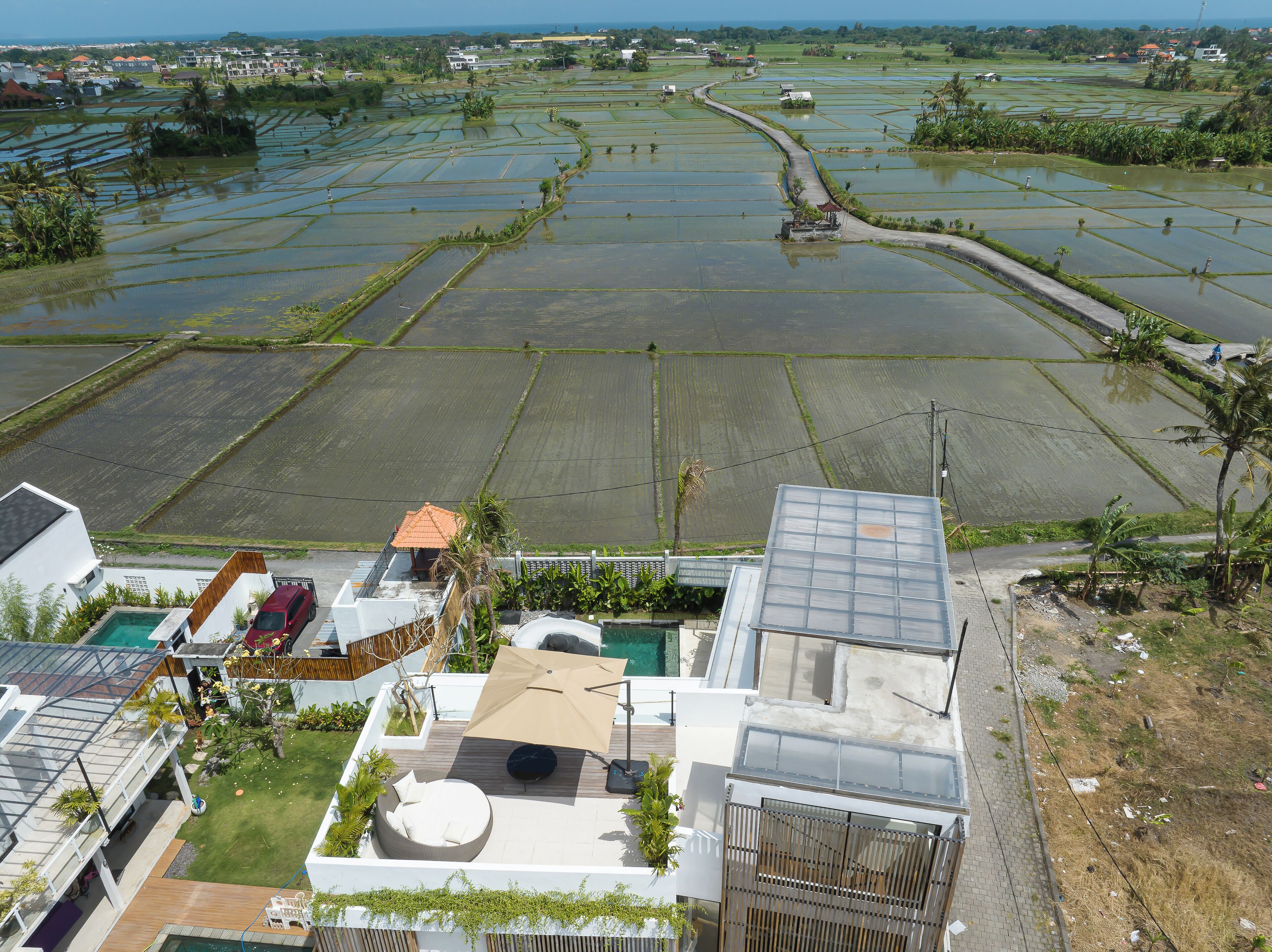 A residential area with a modern house featuring a rooftop pool, a well-maintained garden, and a view of expansive rice paddies in the background.