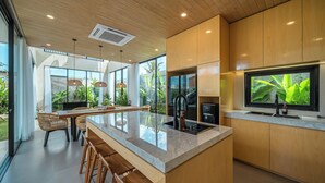 A modern kitchen with a large island, wooden cabinets, and a large window overlooking greenery. - Villa Lumira (Tabanan)