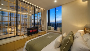 A modern hotel room with a large window, a bed with multiple pillows, a bedside table with a TV displaying "NETFLIX", and a view of the cityscape at night. - Villa Lumira (Tabanan)