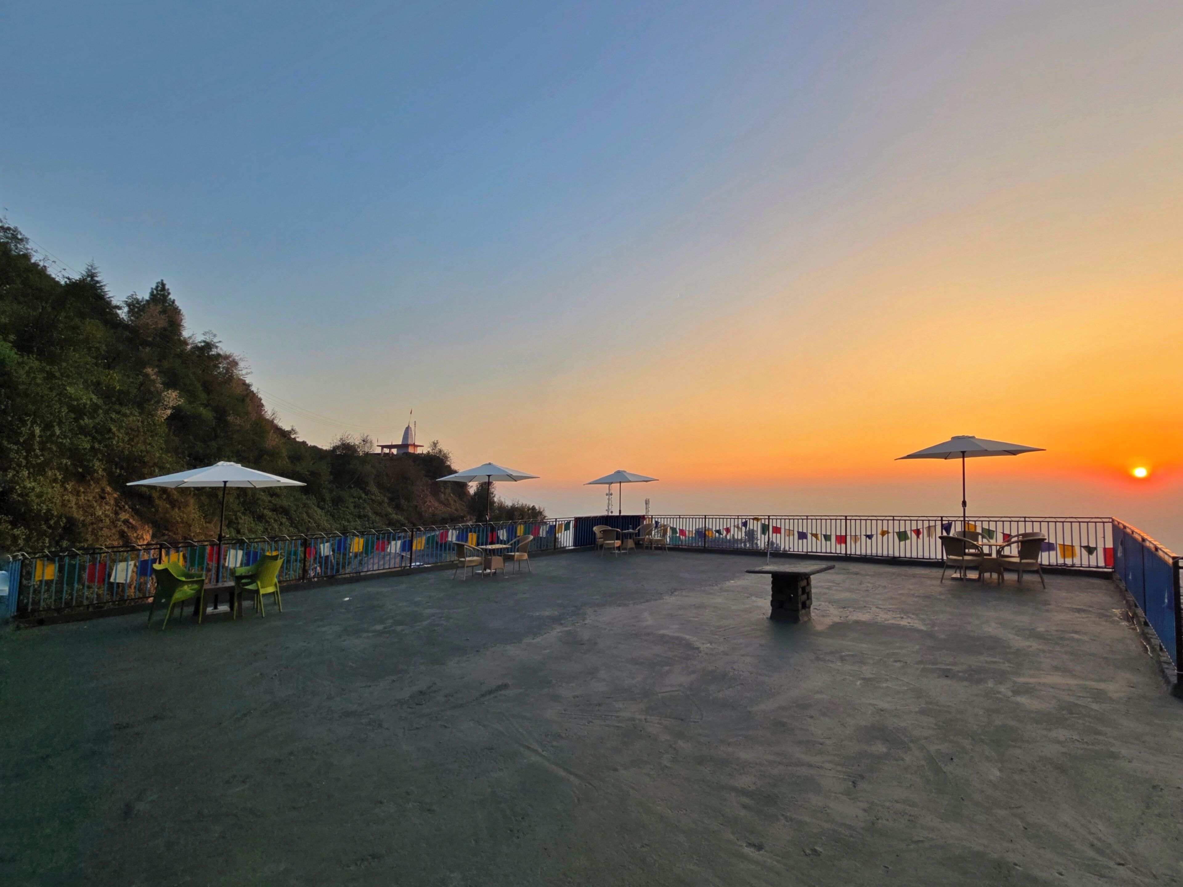 An outdoor seating area with tables and chairs, a flag display, and a sunset view.