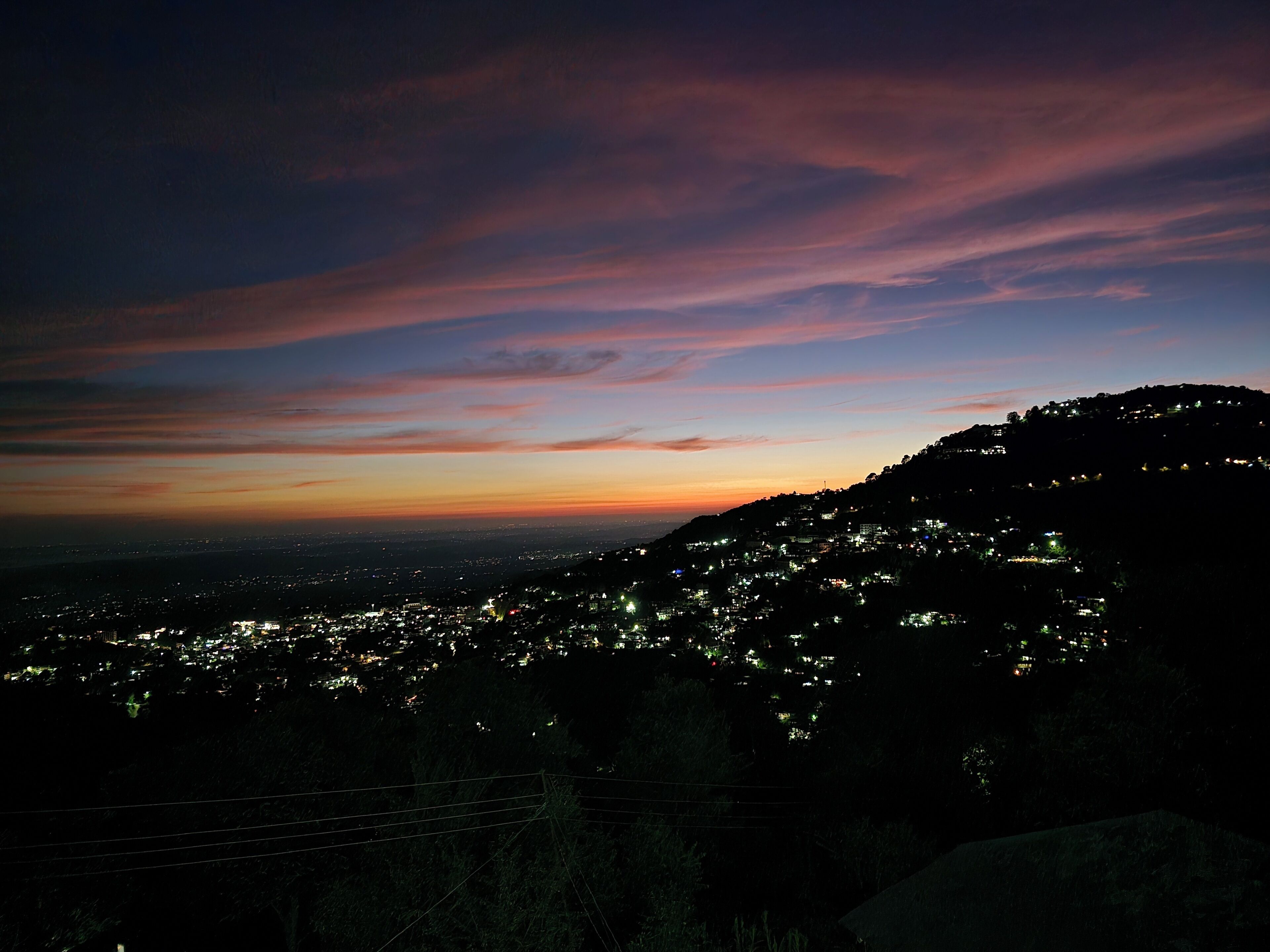 A nighttime cityscape with a sunset sky, illuminated buildings, and a hilly terrain.