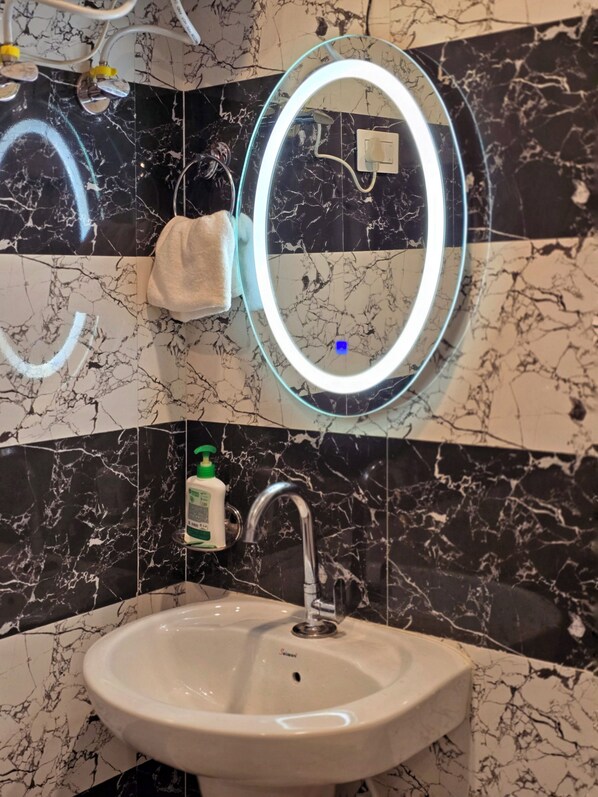 A bathroom with a marble-tiled wall, a white sink, a handheld showerhead, a towel, and a mirror with a light. - Aangan Royal Stay - Your Mountain Home in Dharamshala (Dharamshala)