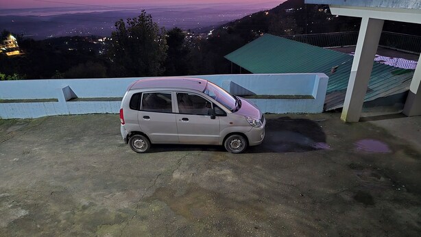 A small car parked on a concrete surface with a view of a distant cityscape at dusk. - Aangan Royal Stay - Your Mountain Home in Dharamshala (Dharamshala)