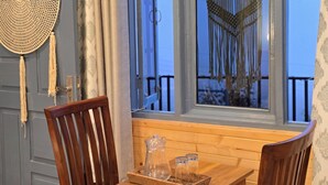 Un tavolo in legno con una brocca di vetro trasparente e due bicchieri, una sedia in legno, e vista sull'esterno attraverso una finestra con decorazione in macramè.