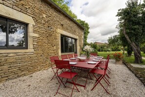 Un espace extérieur aménagé pour les repas, avec un bâtiment en pierre, une terrasse en gravier et des chaises et des tables rouges.