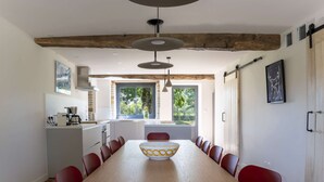 A dining area with a long wooden table, red chairs, and a bowl in the center. There are pendant lights above the table, and a kitchen with a stove and sink is visible to the left.