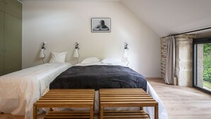 A bedroom with a large bed, two bedside tables, a wooden bench, a picture on the wall, and a window with a view of greenery.