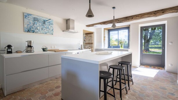 A modern kitchen with a central island, bar stools, a coffee machine, and a large window providing natural light.