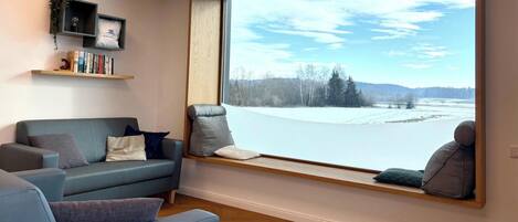 A cozy living room with a large window, a blue sofa, and a bookshelf with books and decorative items.