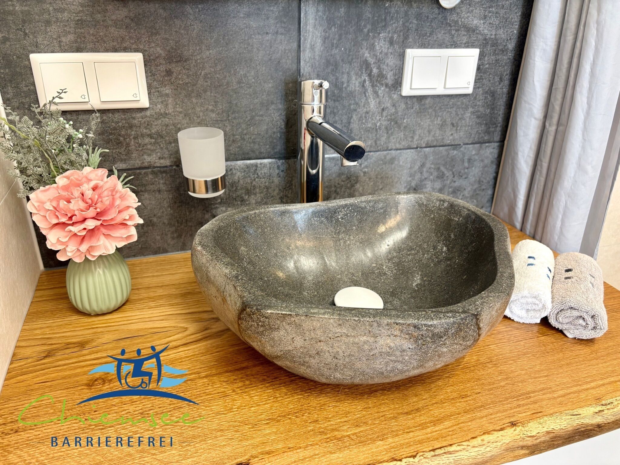 A stone sink with a soap bar, a vase with flowers, and rolled towels on a wooden countertop.