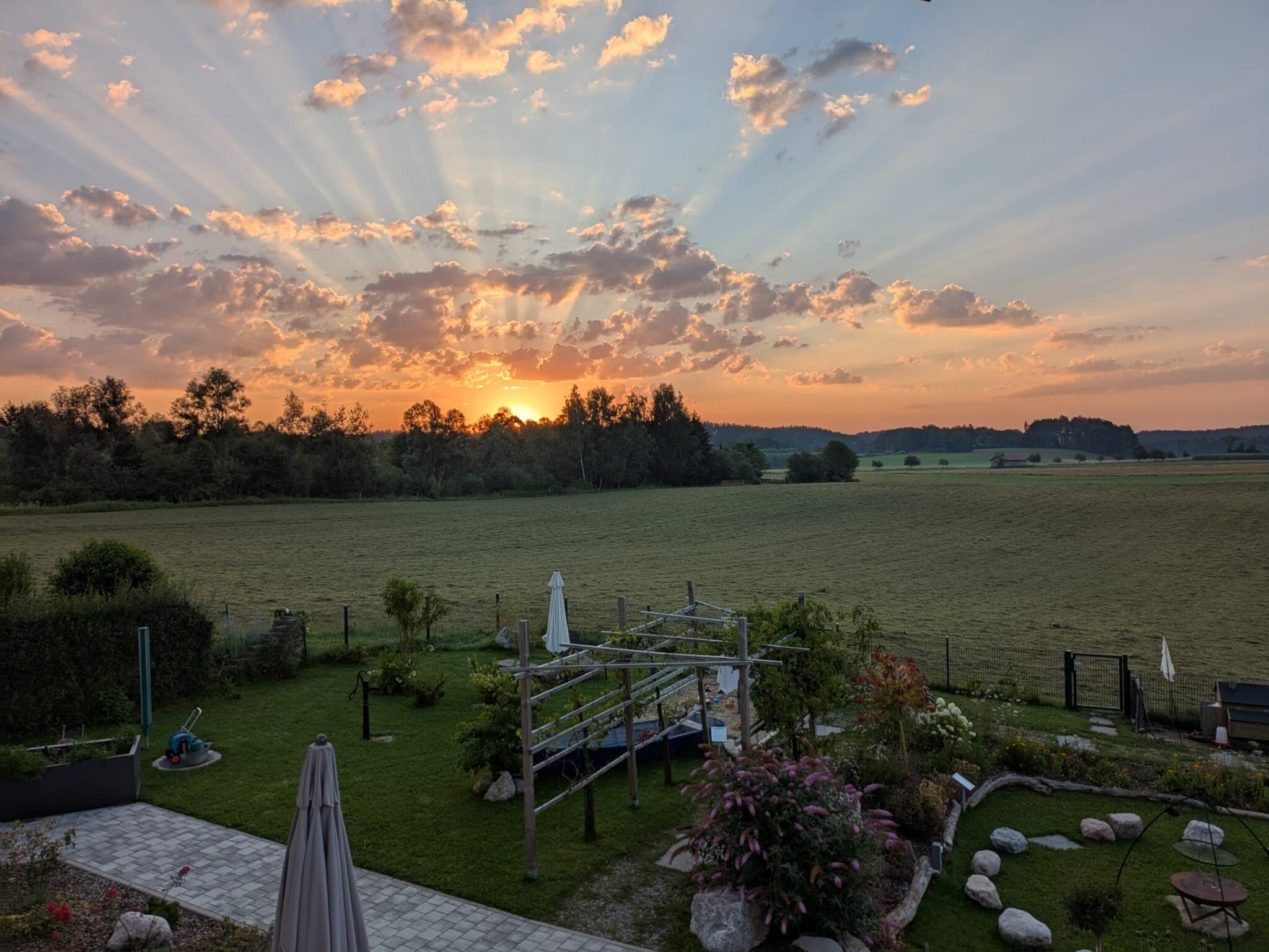 Ein Garten mit einer Pergola, einer Rasenfläche und Blick auf einen Sonnenuntergang über einem Feld.