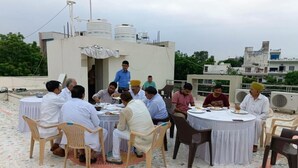 Eine Gruppe von Personen sitzt an runden Tischen auf einer Dachterrasse mit Fliesenboden, umgeben von Gebäuden und Bäumen.