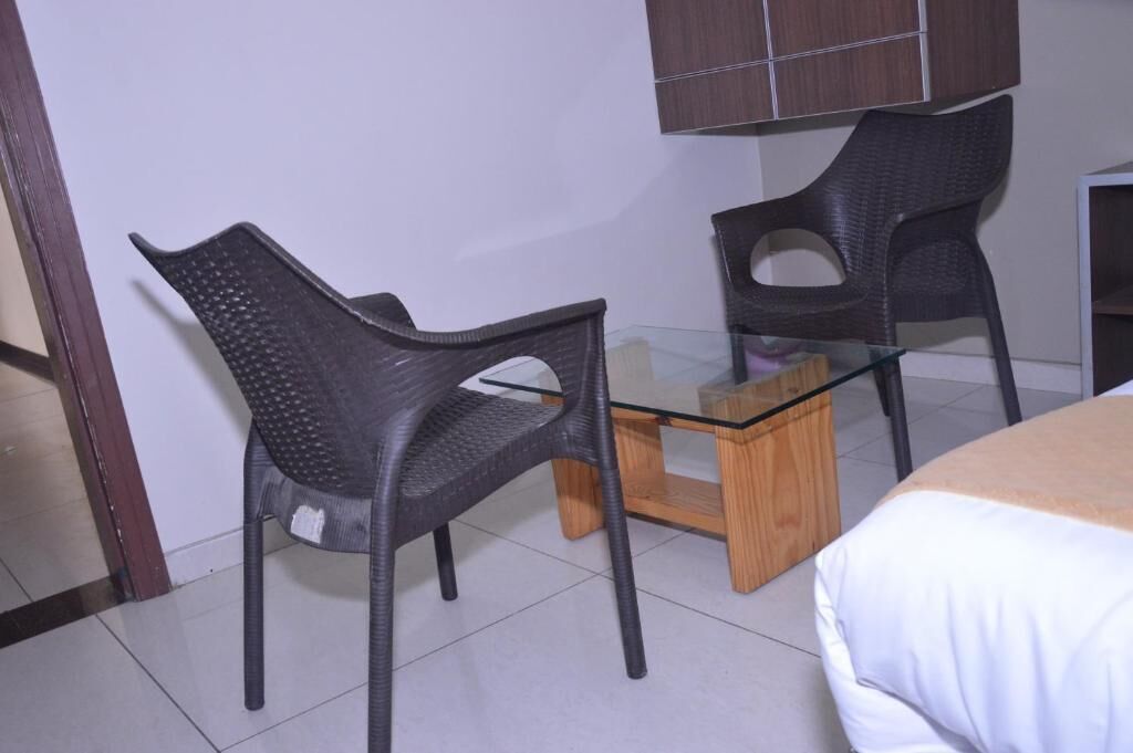 A room with two black plastic chairs, a glass-top table, and a bed with a white cover.