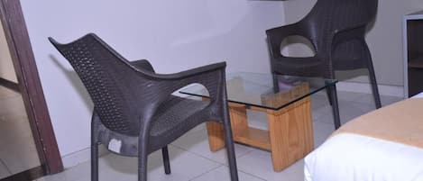 A room with two black plastic chairs, a glass-top table, and a bed with a white cover.