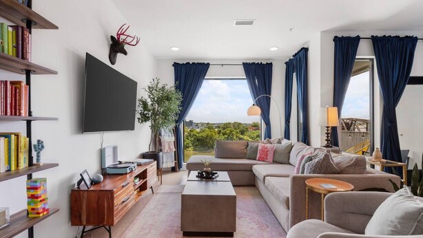 A modern living room with a flat-screen TV, a bookshelf, a coffee table, a sofa, and a view of greenery outside.