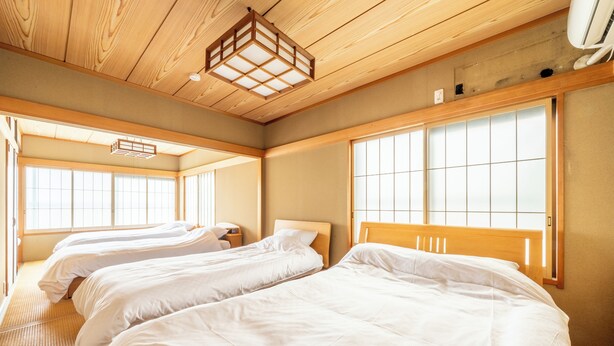 Camera d'albergo con tre letti, soffitto in legno, ampie finestre e una tradizionale lanterna in stile giapponese.