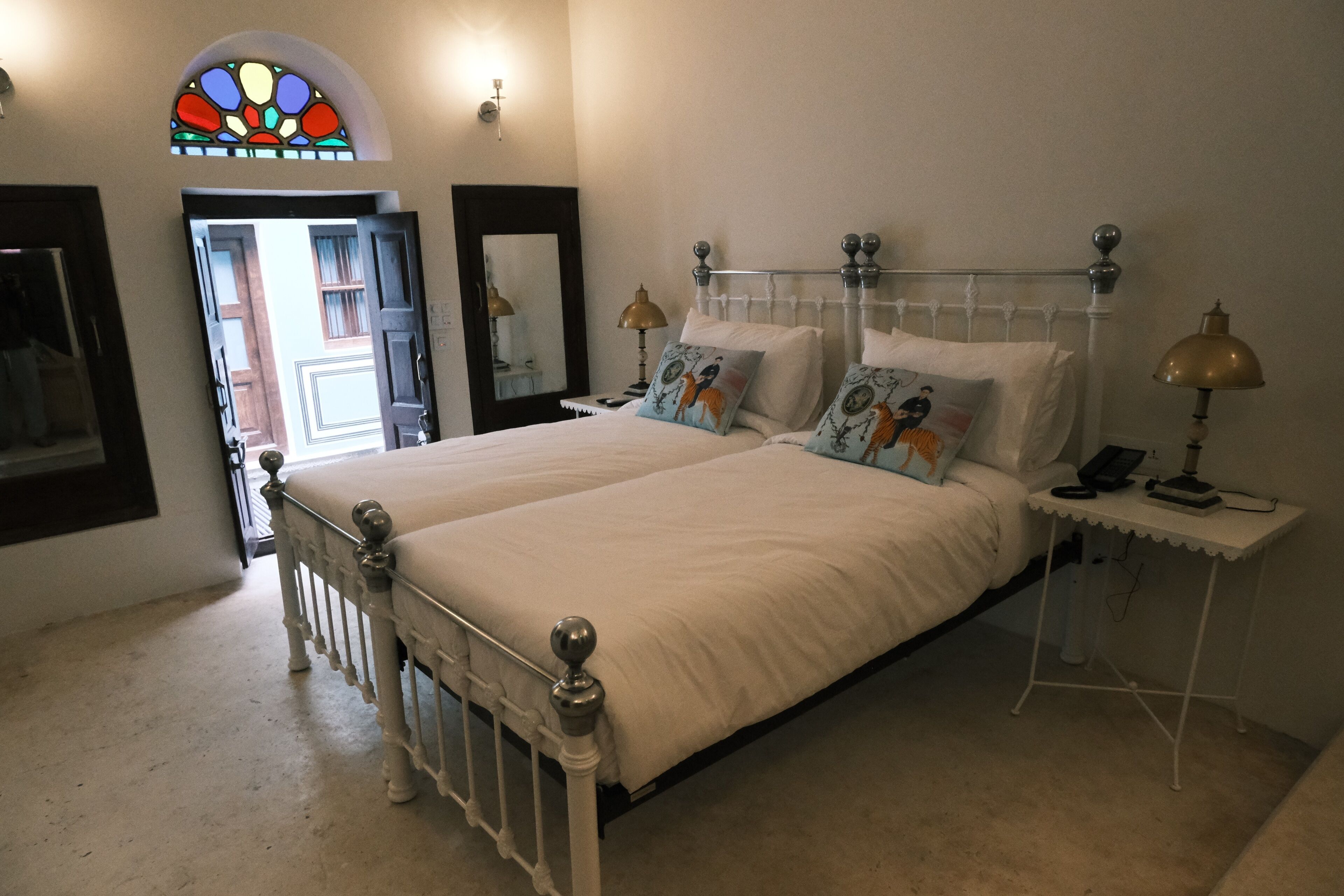 Een slaapkamer met een groot bed, een nachtkastje met een lamp, een spiegel en een glas-in-loodraam.