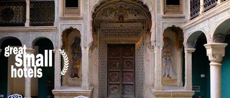 An ornate building with a large central door, multiple arches, and a courtyard with white metal chairs and tables.