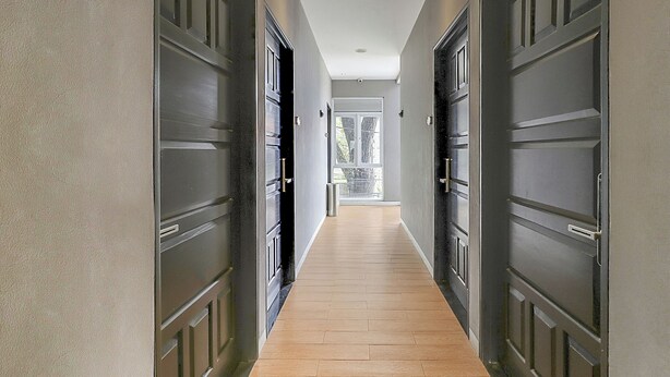 A hallway with dark wooden doors and a light wooden floor, leading to a bright room with a window. - Capital O 94727 Regia Cihampelas Ciwalk (Bandung)