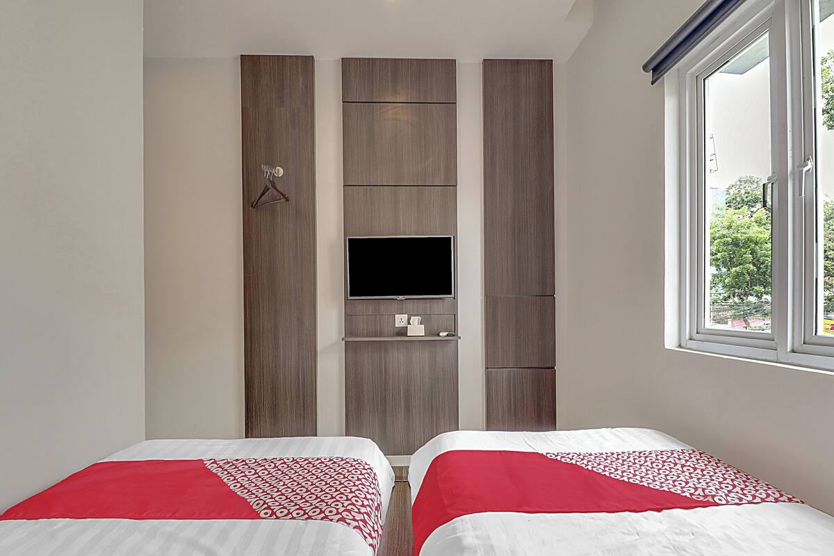 A bedroom with two beds, a TV mounted on a wooden panel wall, and a window with a view of greenery.