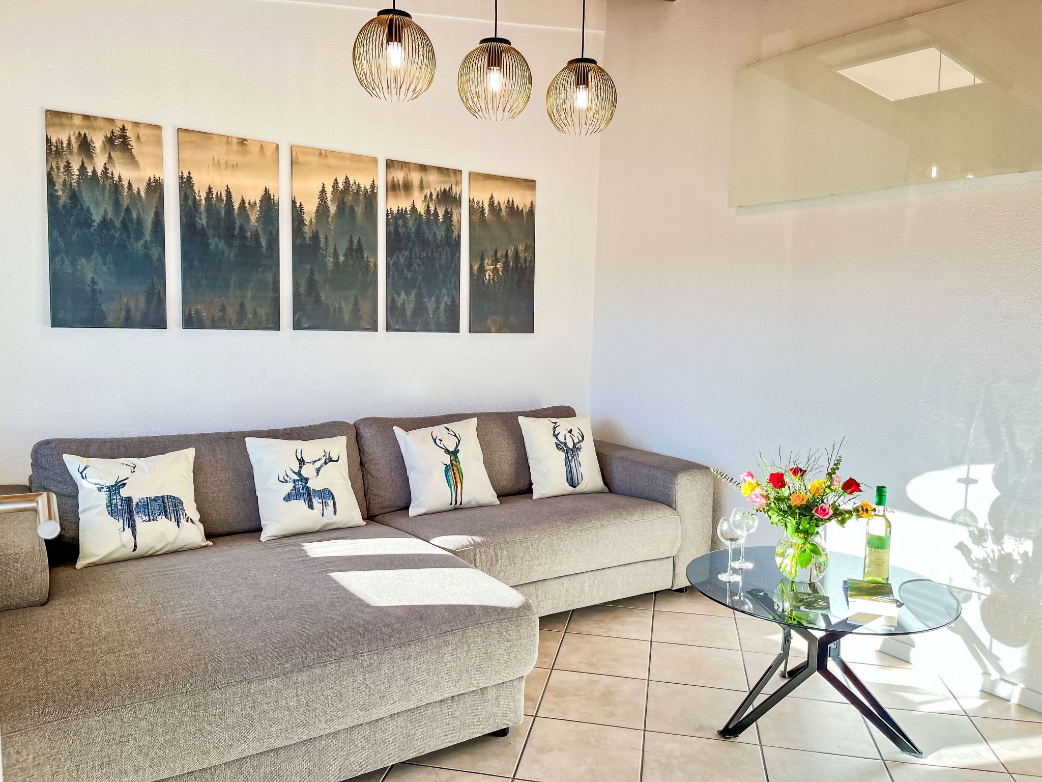A modern living room with a grey sofa, four wall-mounted paintings of forests, and a glass coffee table with a floral arrangement.