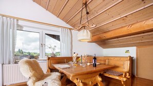 Een houten eettafel met banken, een witte, pluizige stoel, een hanglamp en een raam met gordijnen.