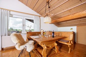 Une table à manger en bois avec des bancs, une chaise blanche et moelleuse, un luminaire suspendu et une fenêtre avec des rideaux.