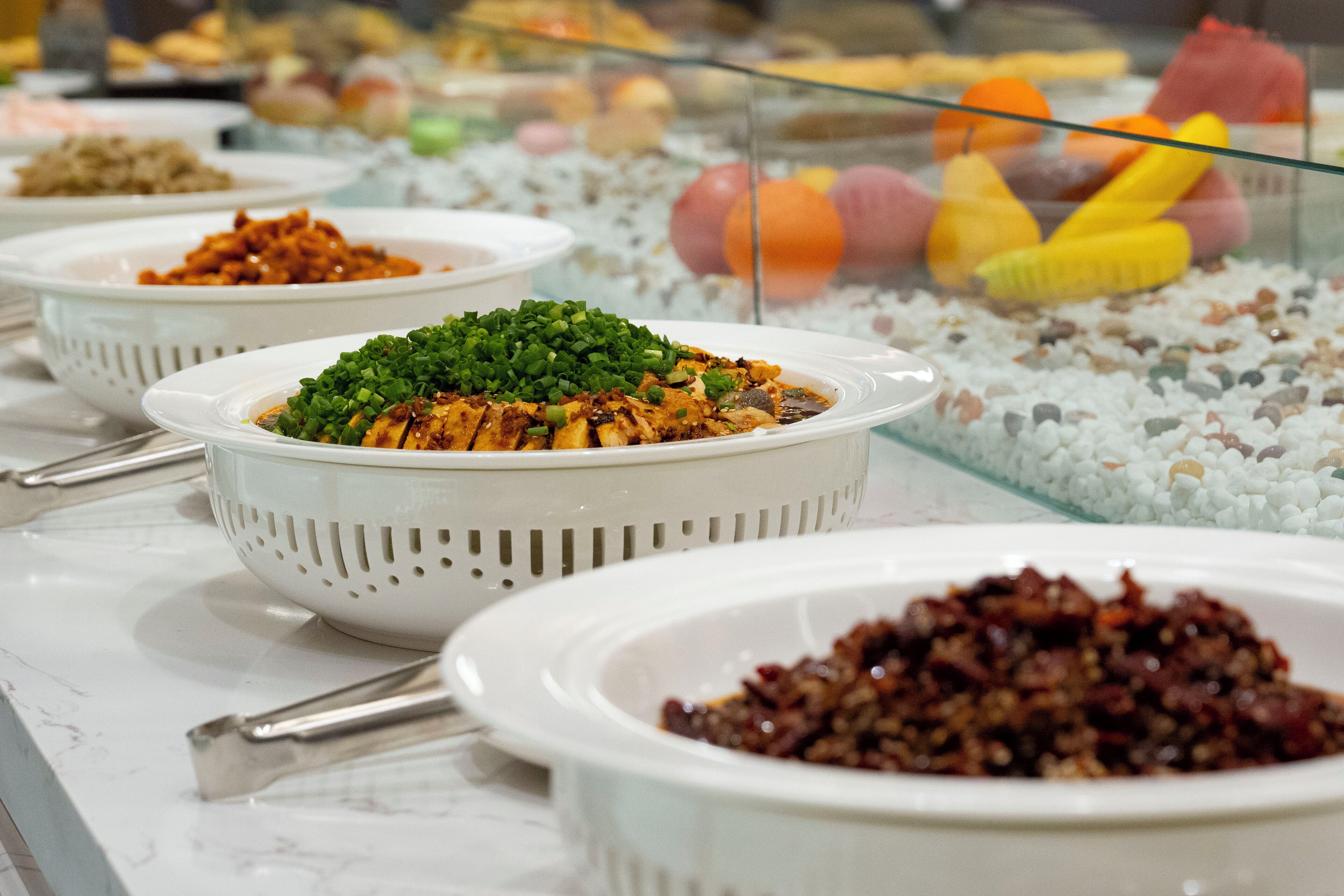 A buffet with various dishes, including a bowl of green vegetables and a bowl of red sauce.