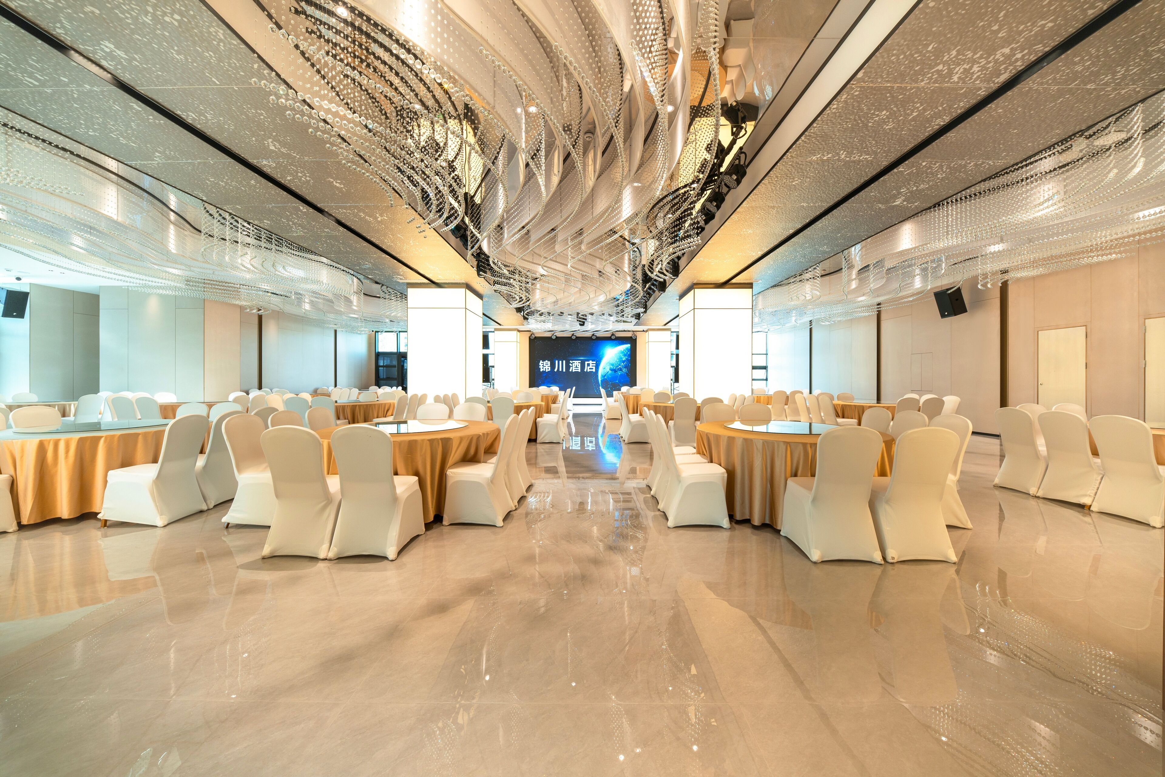 Una amplia sala de conferencias con mesas redondas y sillas blancas, una pantalla grande al frente y un diseño de techo moderno.