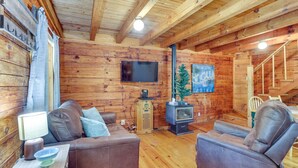 A cozy log cabin interior with two leather sofas, a wooden TV stand, a small heater, and a staircase.