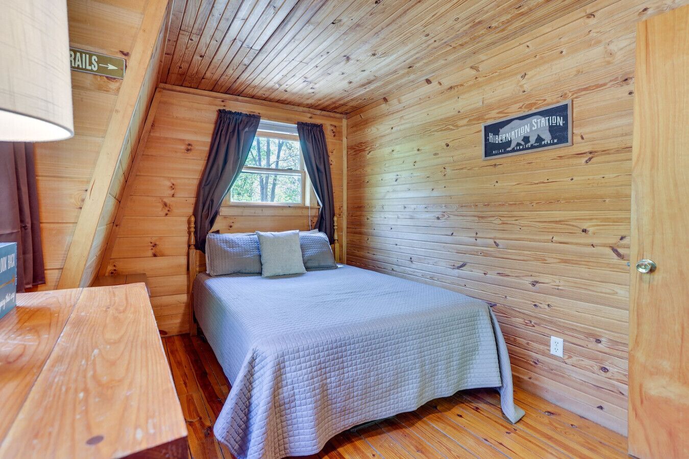 Een slaapkamer in een houten blokhut met een bed, een nachtkastje, een raam met gordijnen en een bordje met de tekst 'winter slaapverblijf'.