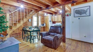 Interior rústico de una cabaña con una escalera de madera, un comedor con mesa y sillas, un sillón de cuero y una zona de cocina con armarios y electrodomésticos.