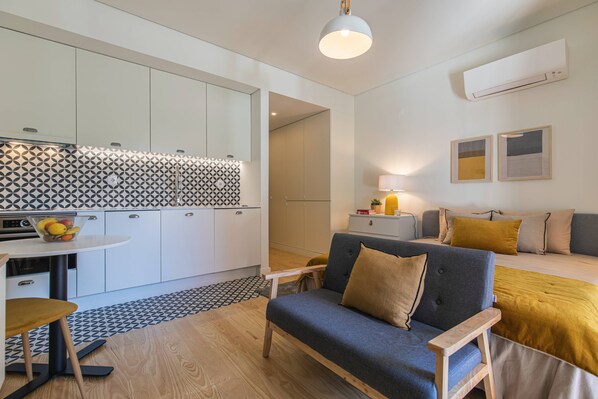 A modern kitchen with white cabinets and a patterned backsplash, a dining area with a table and chairs, and a living area with a sofa and cushions.