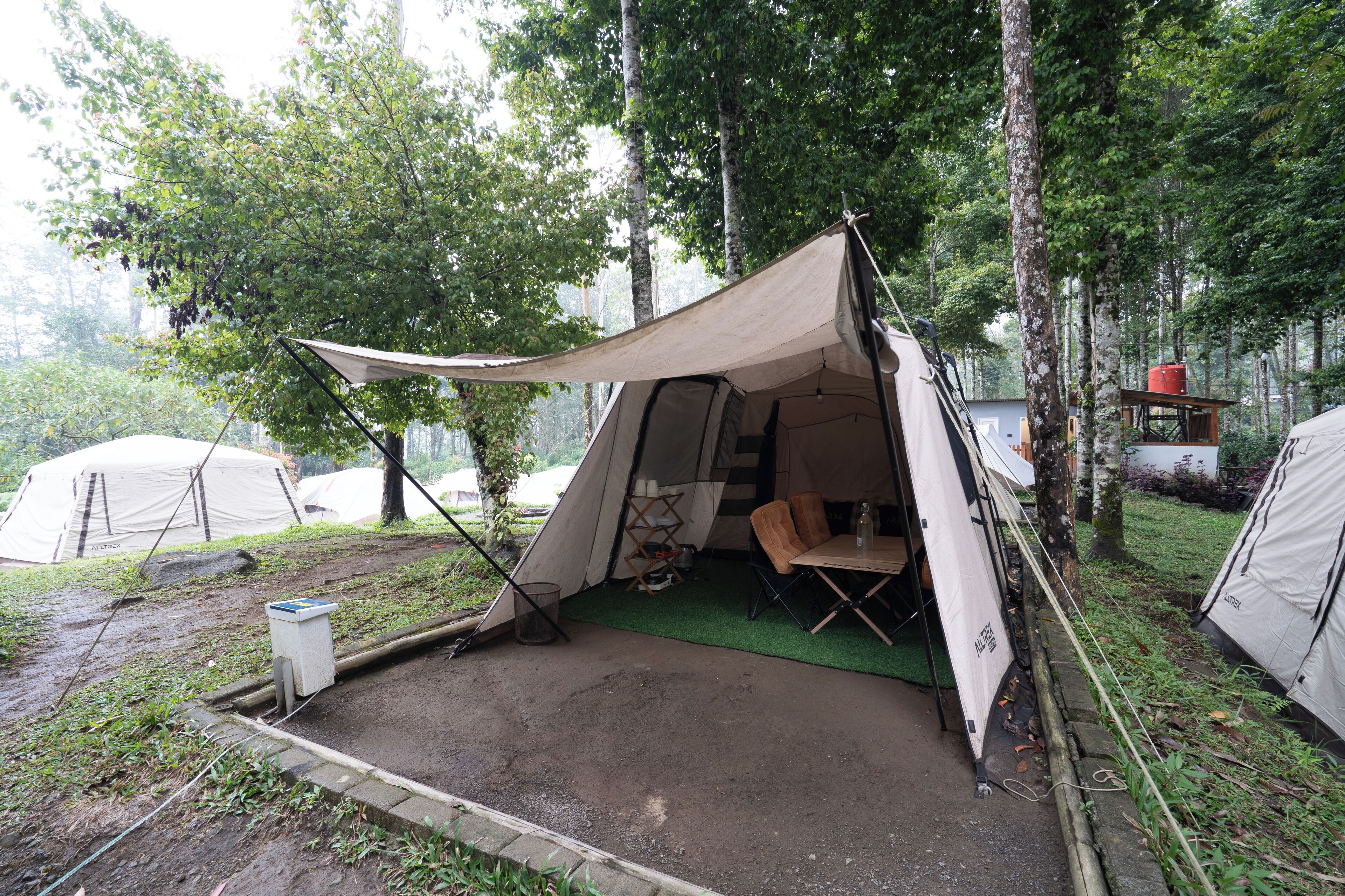 A camping site with a tent set up in a forested area, a small table with chairs, and a water tank.