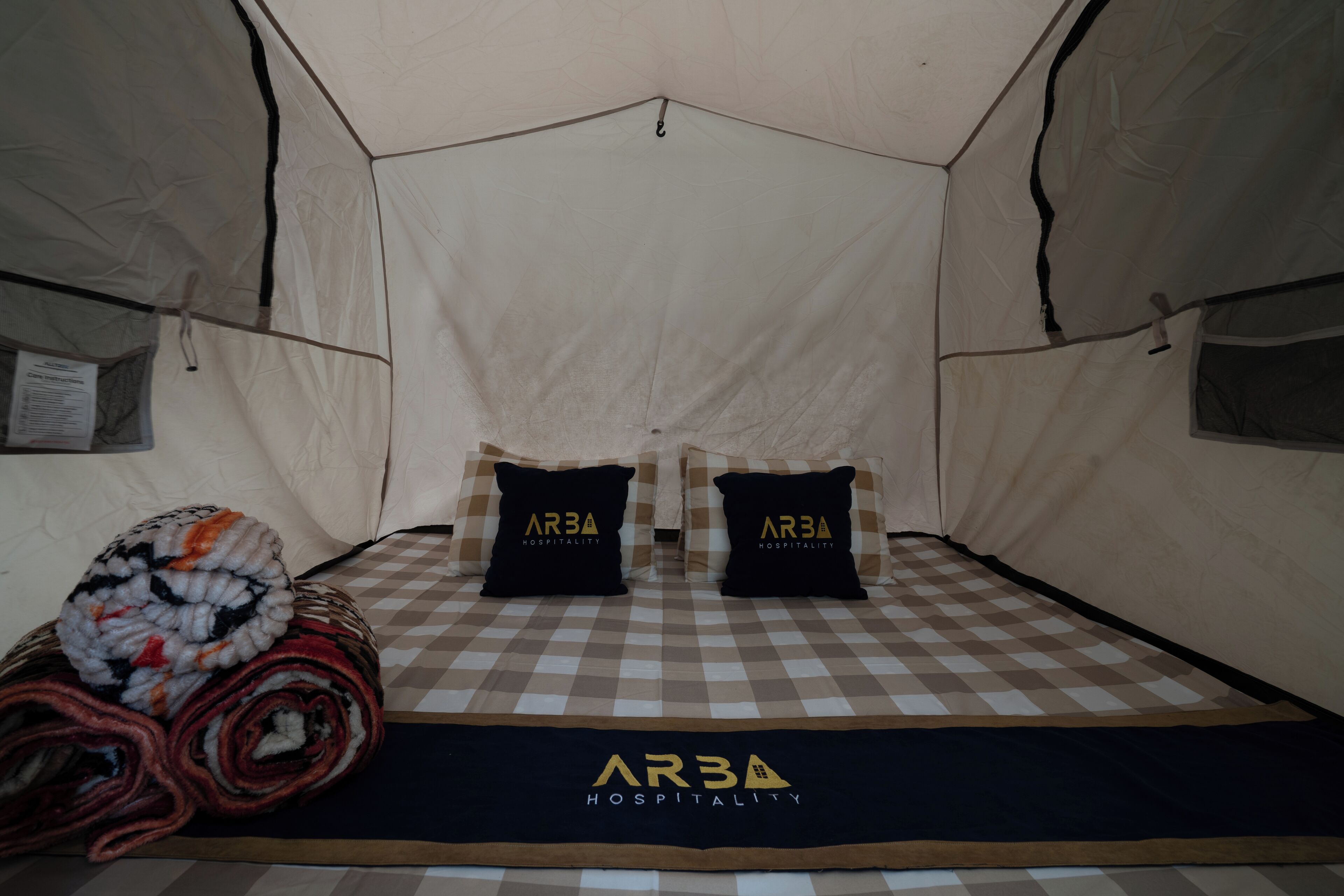 A tented room with two black pillows featuring 'ARBA HOSPITALITY' branding, a checkered floor, and a rolled-up blanket.