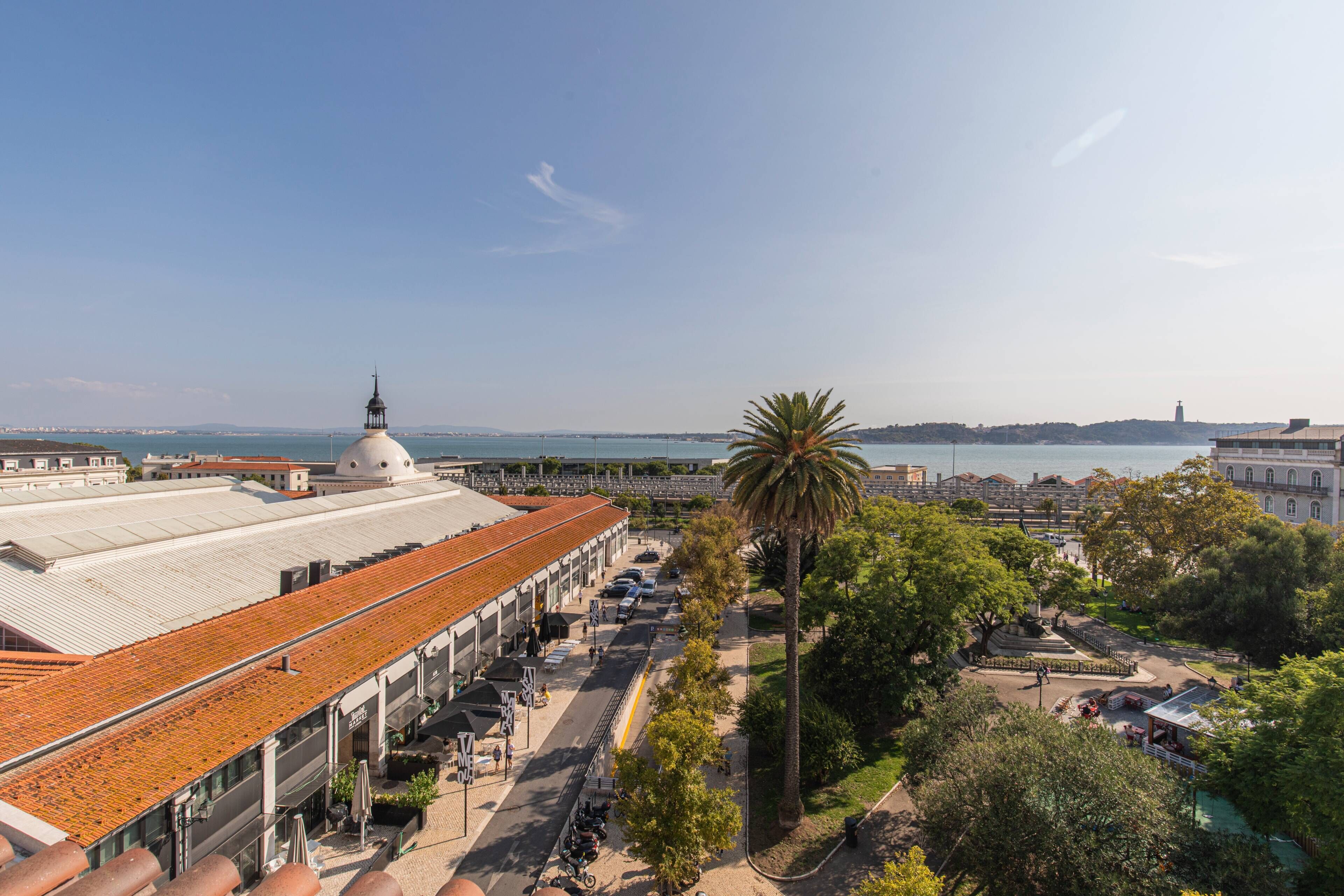 A coastal building with a dome and a red-tiled roof, adjacent to a palm tree and a promenade.