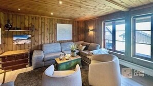 A cozy cabin interior with a wooden ceiling, a sofa, a coffee table, and a large window offering a view of the outside.