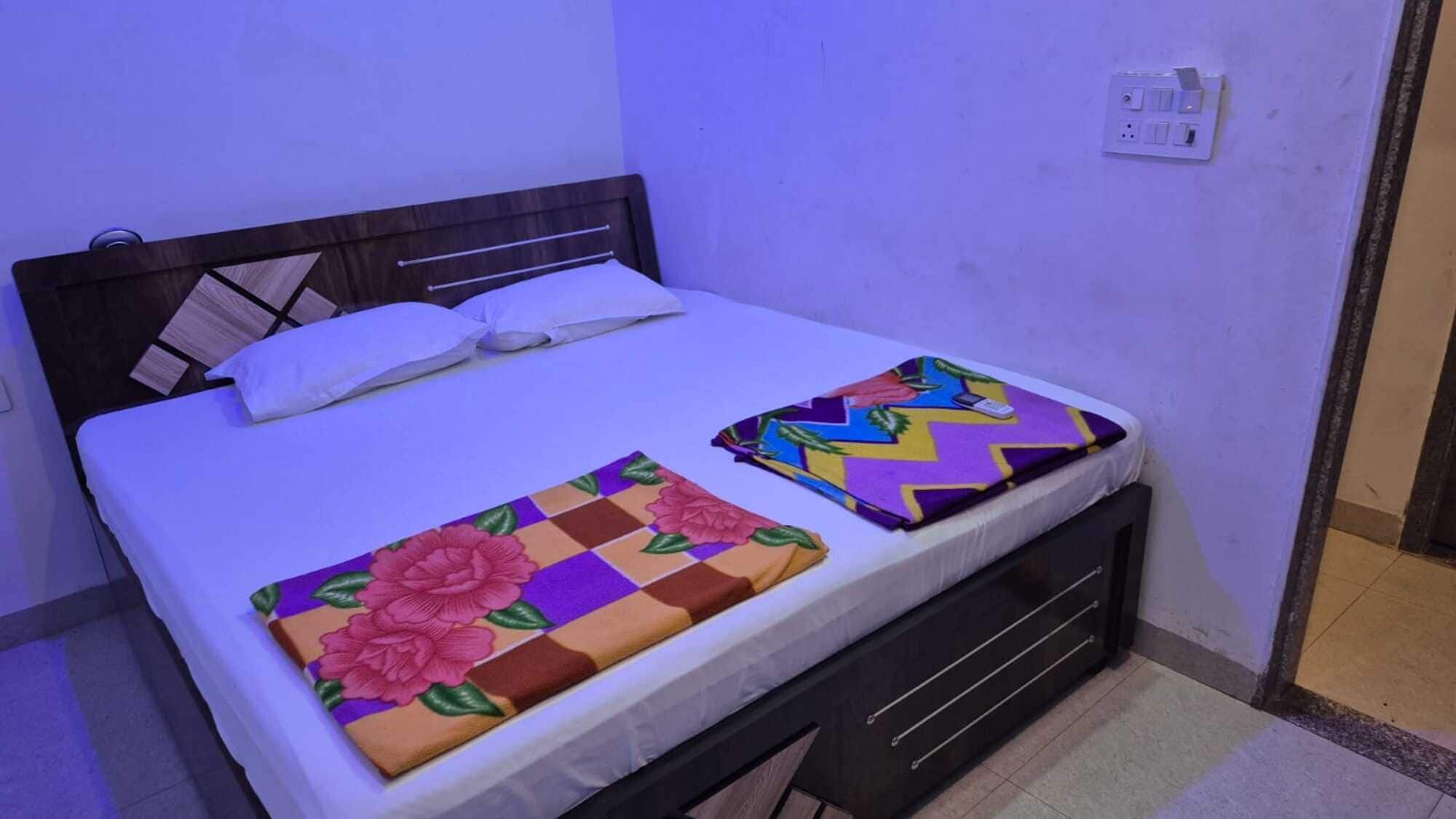 A single bed with a white mattress and a wooden headboard. There are two decorative bedspreads, one with a floral pattern and the other with a zigzag design.