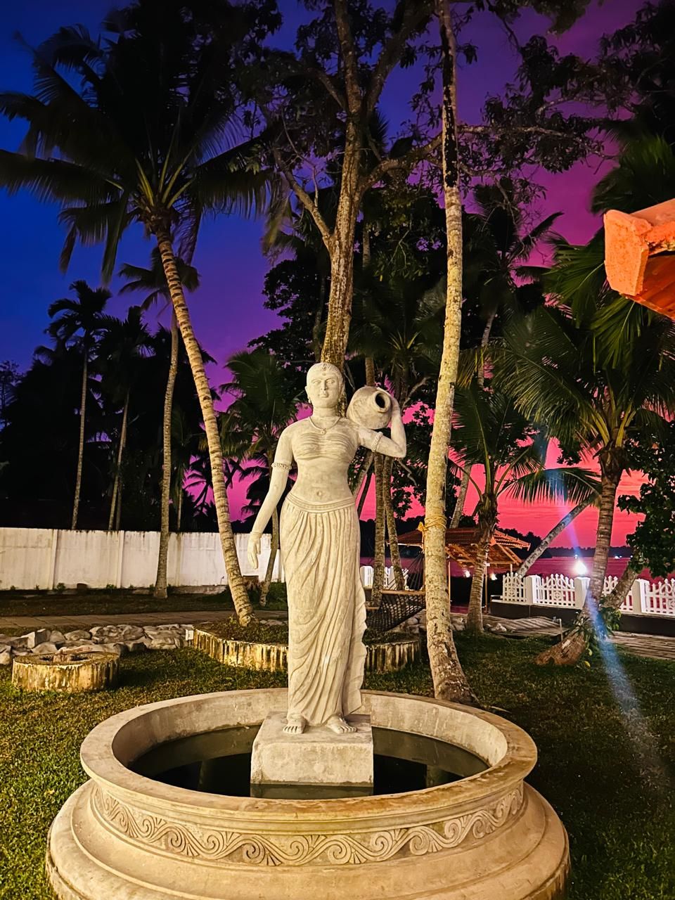 A statue of a woman holding a bowl, surrounded by a decorative fountain, with palm trees and a pink sky in the background.
