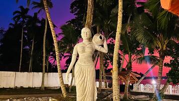 A statue of a woman holding a bowl, surrounded by a decorative fountain, with palm trees and a pink sky in the background.