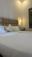 Una cama bien hecha con cabecera y almohadas, una mesita de noche con hervidor, y una lámpara de pared.
