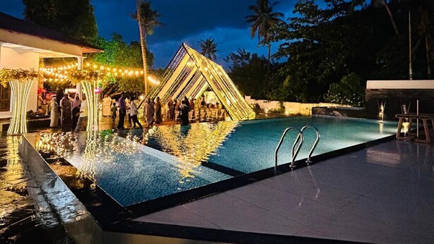 Ein Schwimmbecken mit geometrischer Struktur im Hintergrund, umgeben von Palmen und einem Gebäude mit Lichterketten.
