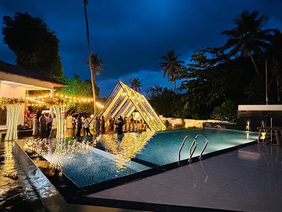 Una piscina con una estructura geométrica al fondo, rodeada de palmeras y un edificio con luces colgantes.
