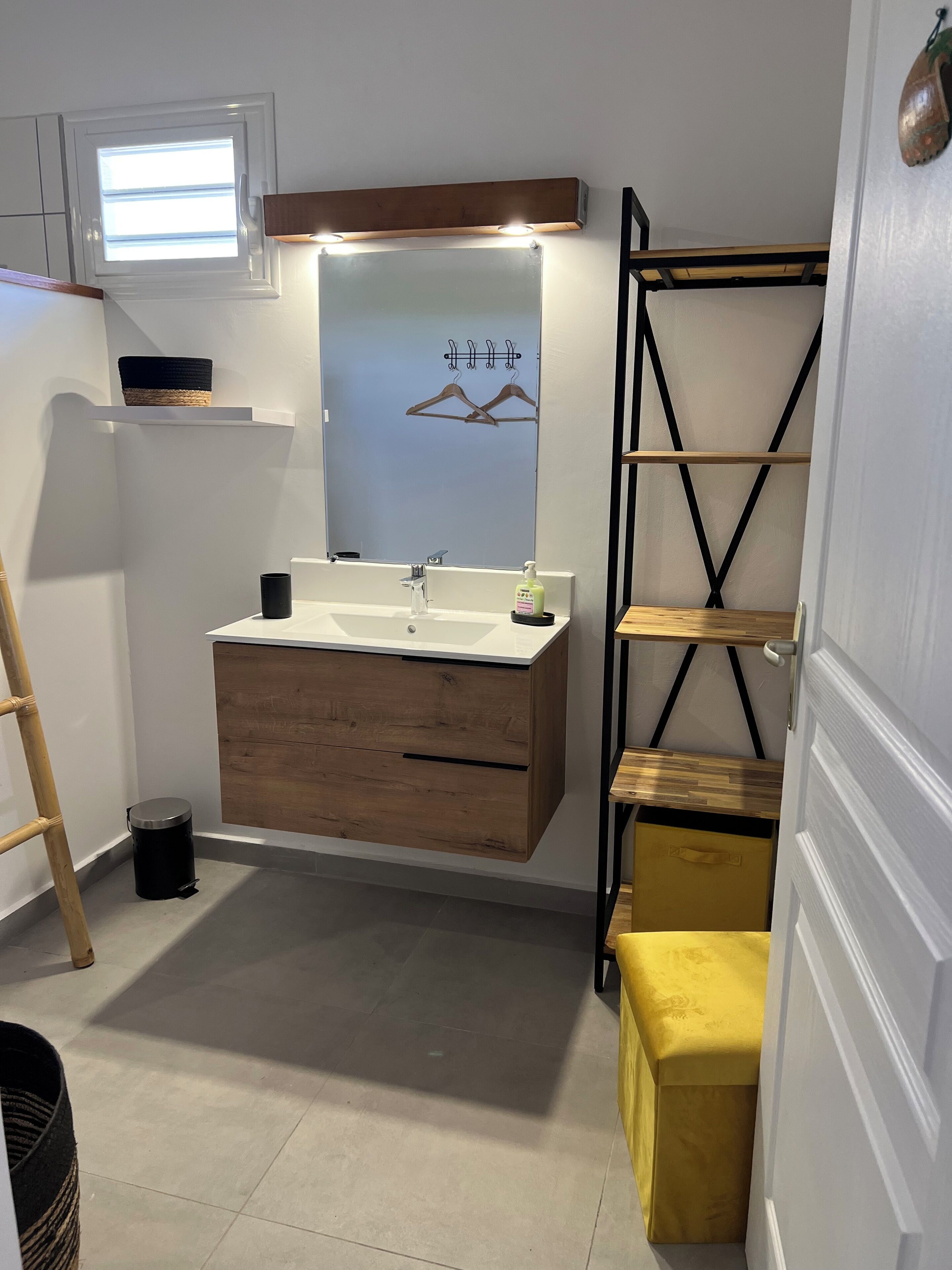 A compact bathroom with a wooden vanity, a mirror with lights, a small shelf, and a yellow bench.
