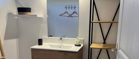 A compact bathroom with a wooden vanity, a mirror with lights, a small shelf, and a yellow bench.