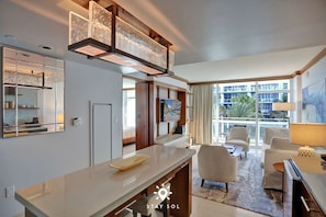 A modern hotel room with a large window, a flat-screen TV, a dining area with a long table, and a seating area with two chairs. - Spectacular Apt - Sea View Luxury & Free Parking (Miami Beach)